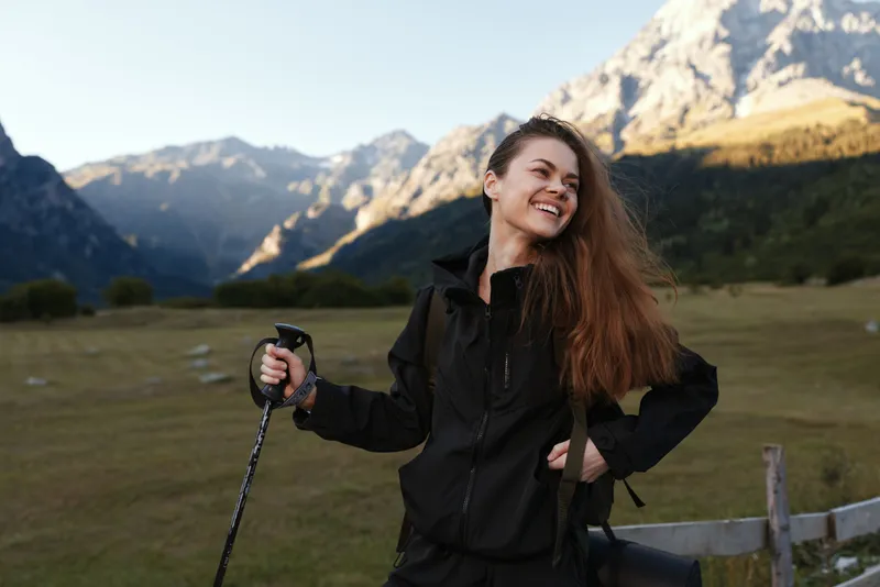 A cheerful woman hiker stands in a wide valley, smiling brightly while gripping a trekking pole. She enjoys a sunny day amid rugged mountains and expansive blue sky.