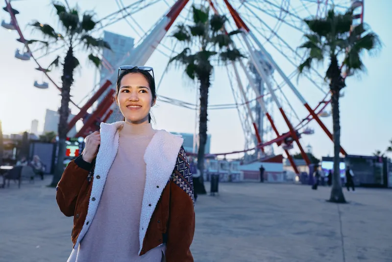 Woman traveler outdoors near a ferris wheel, smiling in a warm coat, enjoying urban leisure, palms sway in the breeze, evening light enhances candid street scene, modern entertainment backdrop for