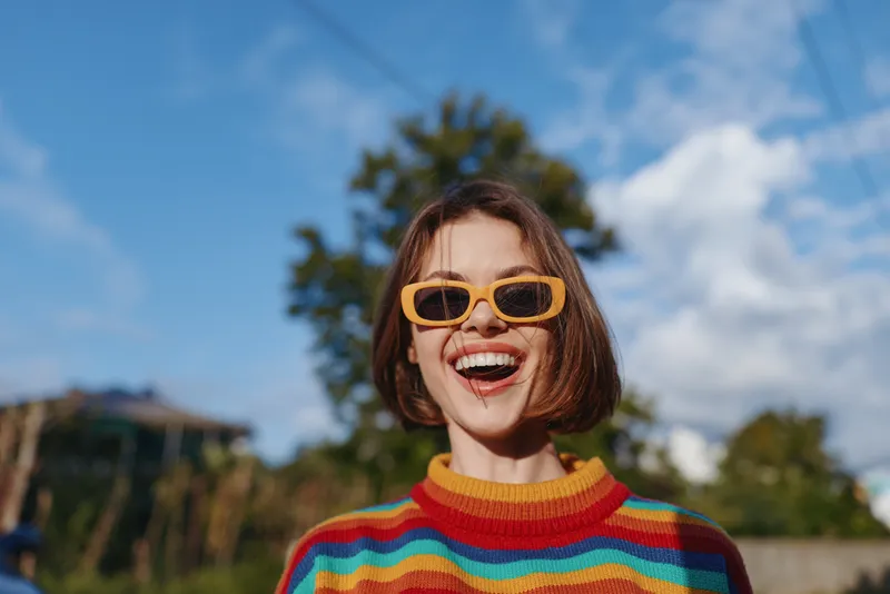 Woman in her 20s smiling in a striped sweater and yellow sunglasses outdoor under a blue sky, short bob haircut, joyful casual outfit, lifestyle portrait for travel fashion