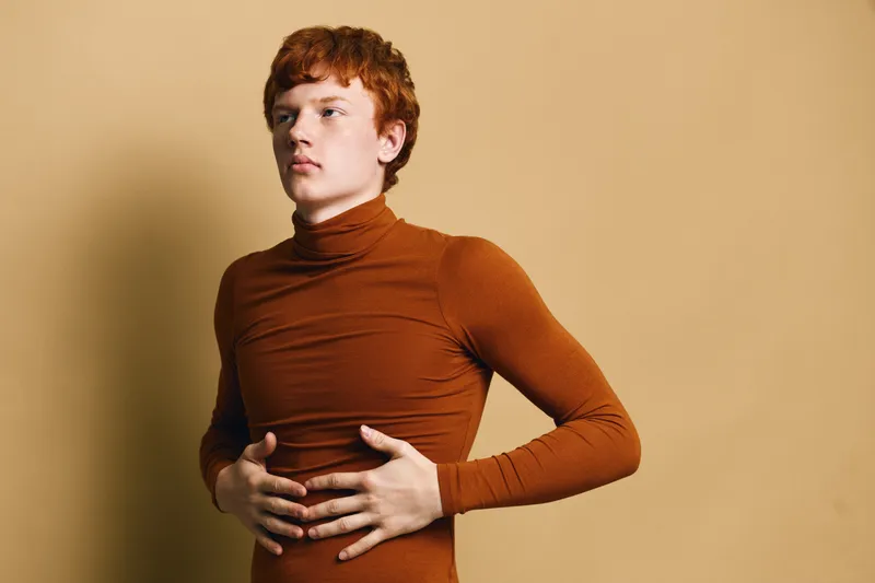 Young man with red hair wearing brown turtleneck standing against beige background holding stomach with both hands looking sideways with serious expression.