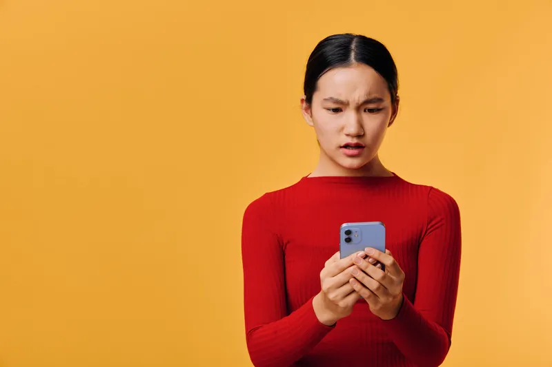 Young woman in red sweater looking confused at smartphone screen with yellow background. Female person holding phone with concern or frustration while reading message or notification.