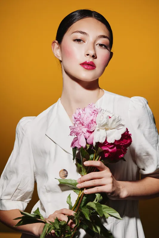Young woman with glossy makeup and vibrant lipstick holds pink and white flowers. She wears a white outfit against a yellow background. Fashion beauty concept with creative styling.