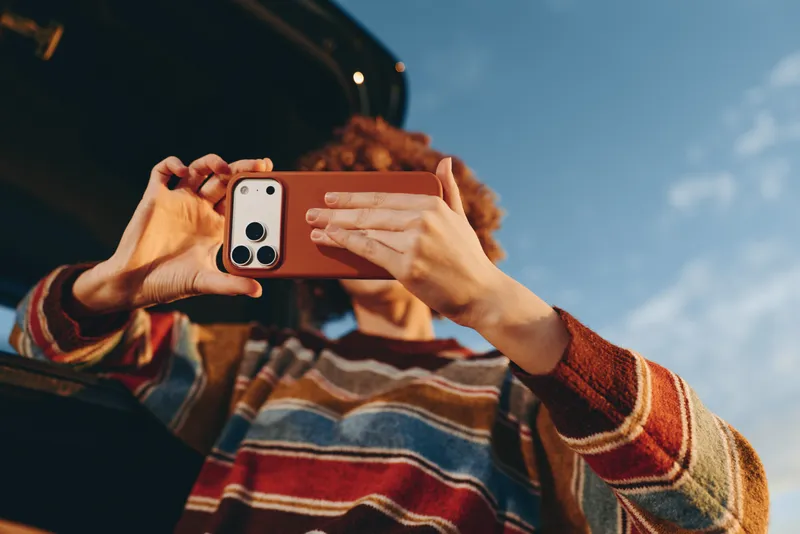 Woman lifestyle outdoors portrait taking smartphone selfie hands raised with smartphone against blue sky curly hair striped sweater warm sunlight candid moment today smile