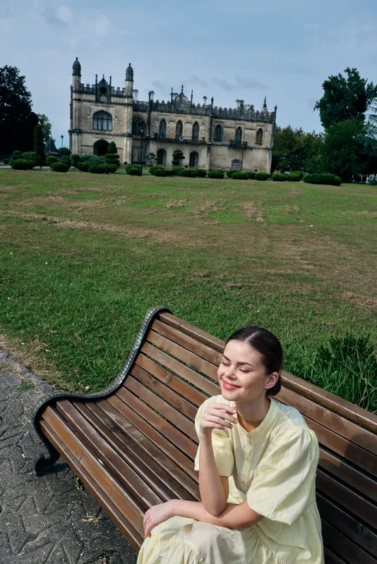 A woman sits on a wooden bench in a lush park, with a historic castle on a distant hill behind her, smile on her face, enjoying a peaceful outdoor moment.