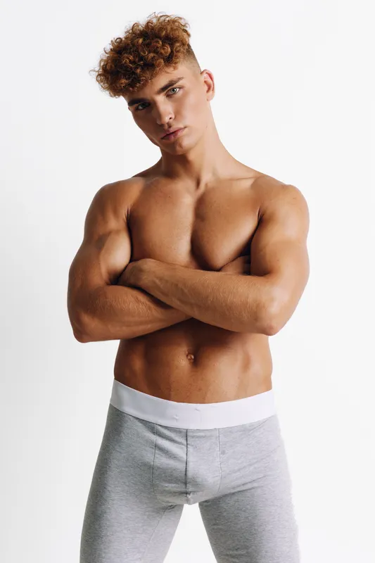 Young man with curly hair standing shirtless with arms crossed wearing gray pants against white background, confident relaxed pose and well-defined muscular body, studio portrait.