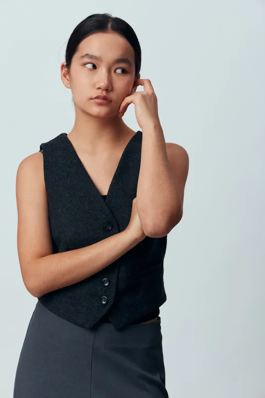 Young Asian woman with dark hair wearing sleeveless vest and skirt stands thoughtful with hand touching face and looking sideways on plain background. Casual confident pose with natural expression.