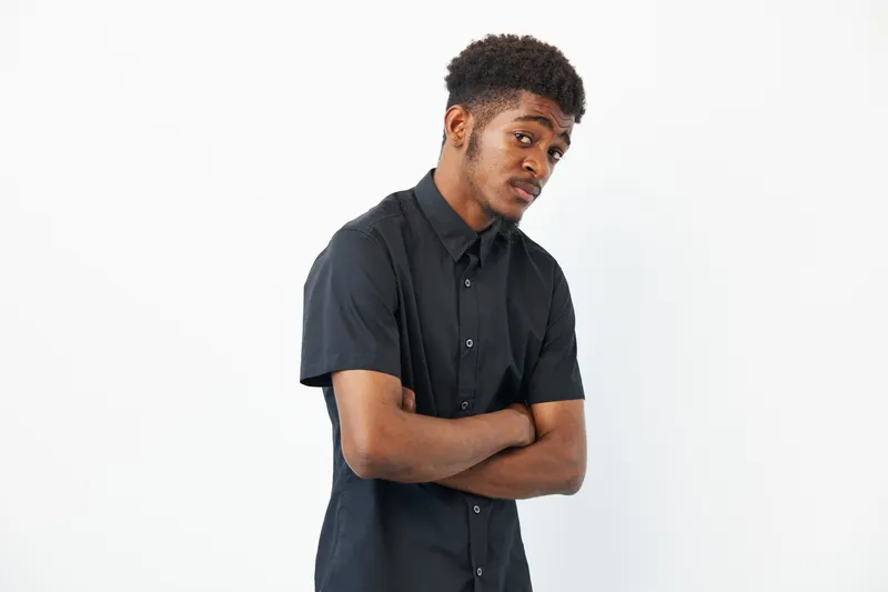 A thoughtful African American man with crossed arms, showcasing a stylish black shirt. His expression embodies contemplation and introspection, set against a clean background.