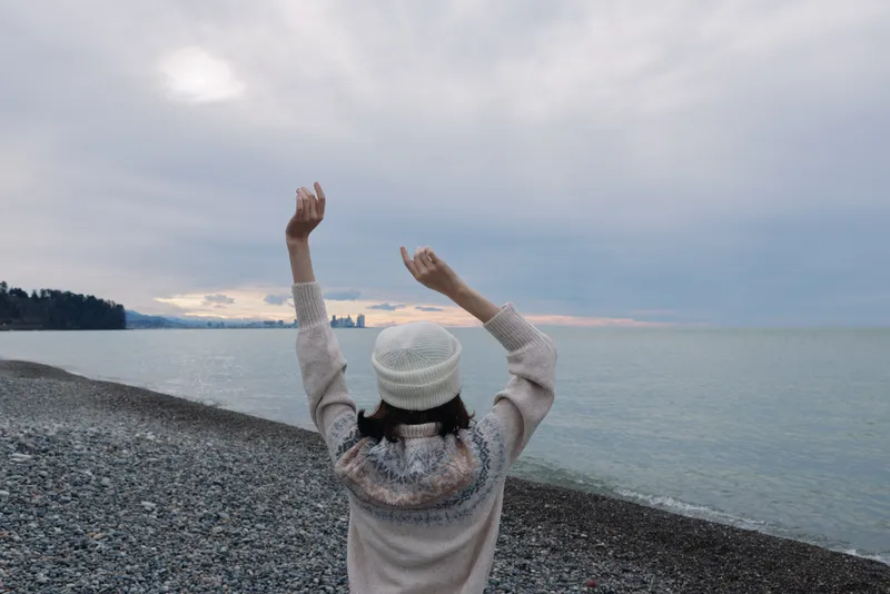 Person stands on a pebble beach with arms raised toward a cloudy sky, wearing a knit sweater and beanie, looking over the sea and horizon for a sense of freedom and serenity