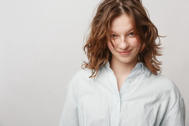 Smiling young woman with curly hair wearing white shirt looks confidently at camera. Studio portrait on light background. Concept of natural beauty, youth, and casual style.