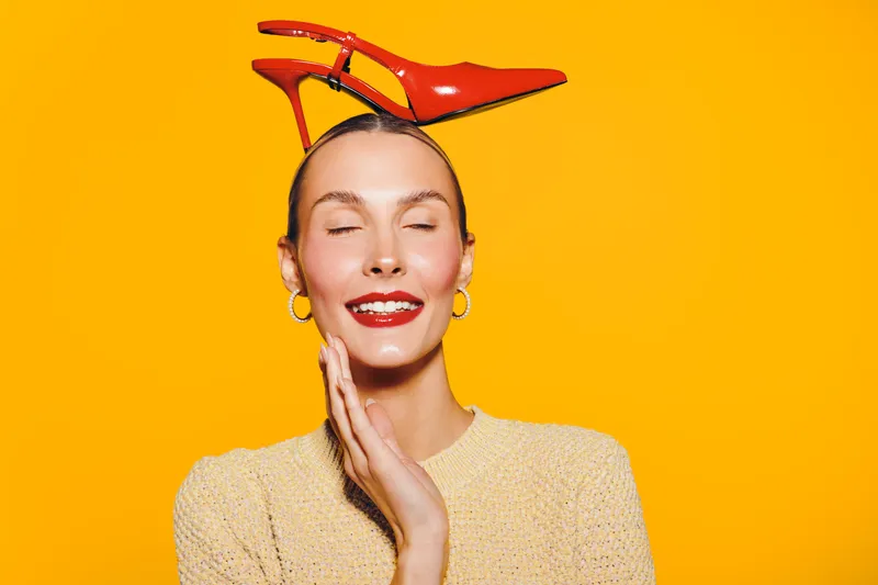 Smiling woman with closed eyes wears red high heel shoe on head against bright yellow background. Concept for fashion, fun, creativity, and unique style portrait in studio.