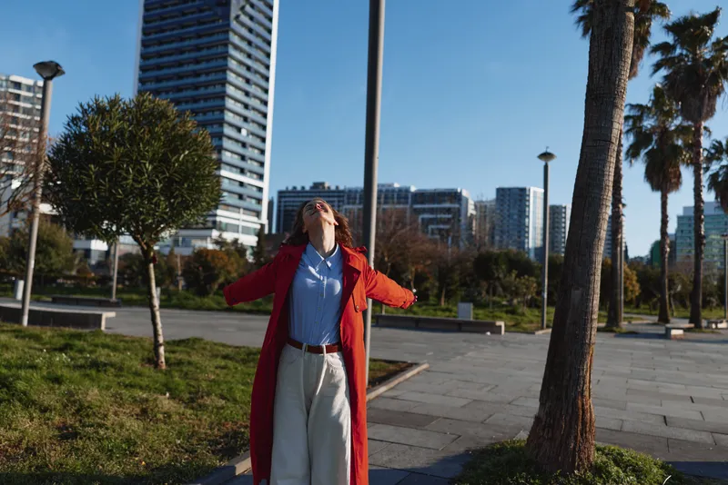 Young woman in a red coat stands with arms outstretched enjoying a sunny urban park with modern buildings in the background and palm trees nearby.