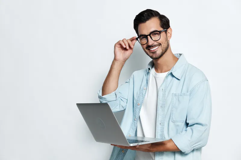 Smiling man wearing glasses holding laptop with hand on chin, expressing confidence and readiness, standing against solid light background. Professional lifestyle concept.