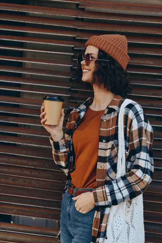Woman with coffee and smile in beanie and plaid shirt on urban street, casual pose conveying authenticity and candid lifestyle with golden hour glow and mindful living mood.
