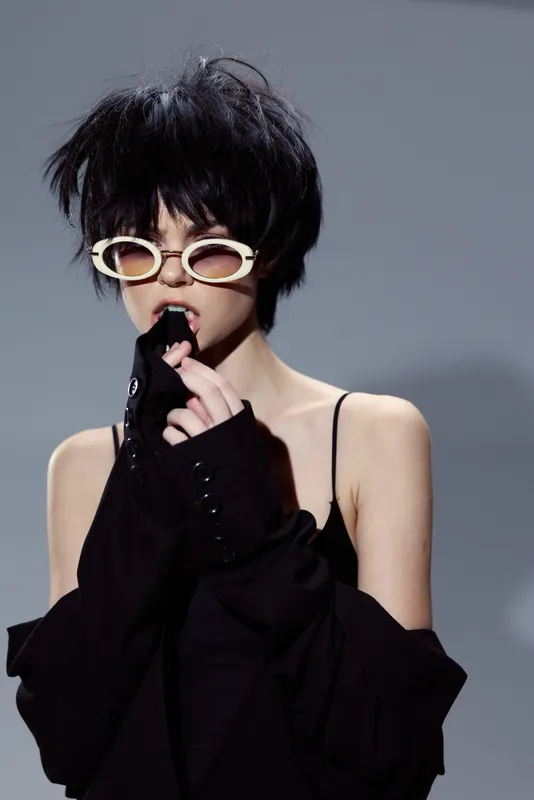 Stylish woman with black hair wearing white sunglasses and black clothing, posing with a mysterious expression against a neutral background.