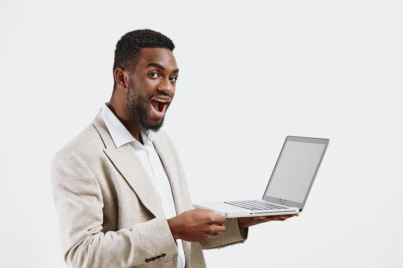 Smiling man holding a laptop computer in hand, wearing a beige blazer and white shirt, looking at camera, isolated on plain white background, studio portrait. People lifestyle concept, cheerful, profe