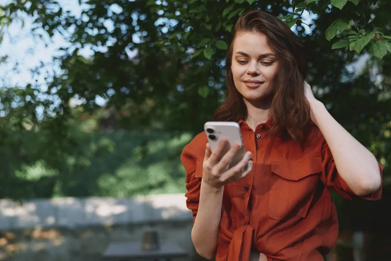 Woman enjoying a casual outdoor moment, looking at her smartphone with a relaxed smile, in a park setting, conveying everyday lifestyle and leisure