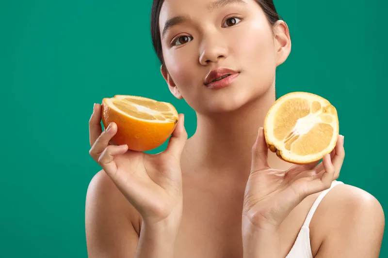 Asian woman holds orange halves toward the camera in a bright studio, showcasing vibrant fruit juice and a fresh, healthy vibe; her warm smile and confident gaze convey energy and wellness.