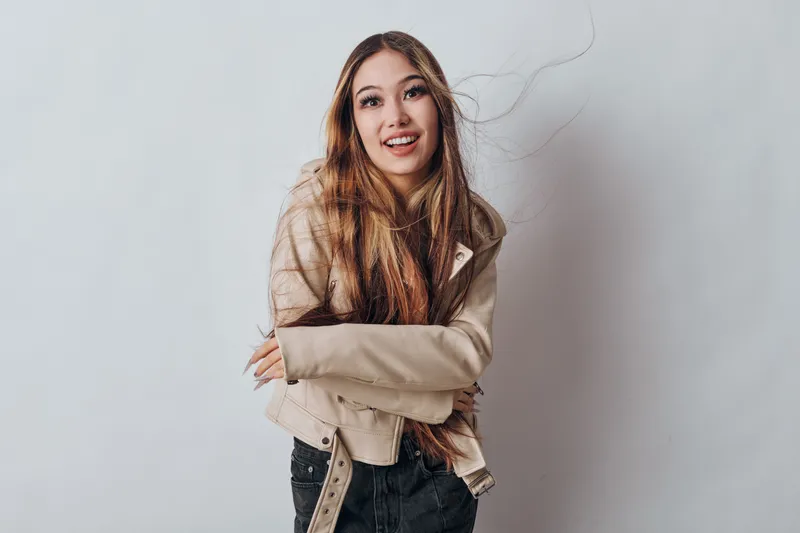 Young woman with long hair and casual jacket smiling and posing against white background in studio. Portrait of happy female with crossed arms and natural look for lifestyle and fashion.