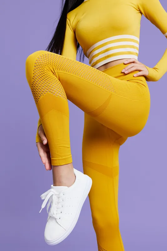 woman sport fitness wearing bright yellow activewear stretching leg in white sneakers against purple background, healthy lifestyle and workout concept