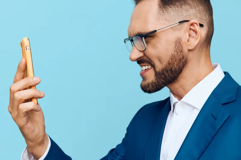 A professional business man smiles while looking at his smartphone against a solid blue backdrop, conveying confidence, focus, and positive emotion.