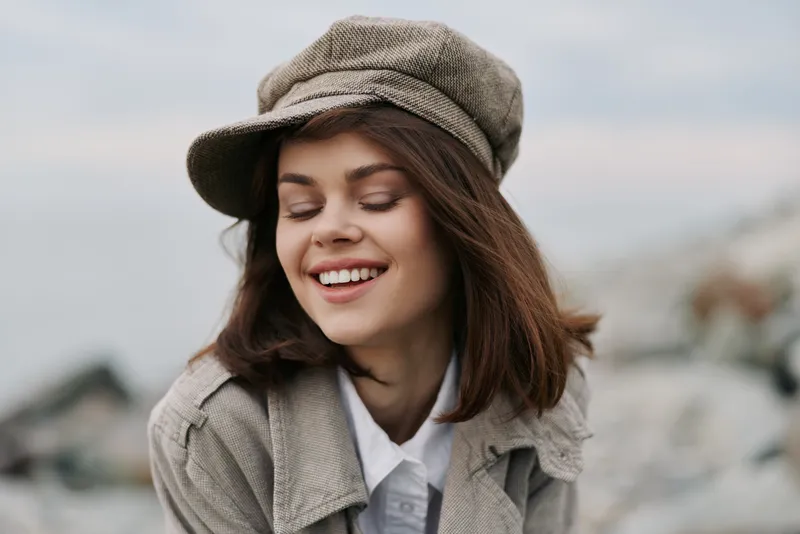 Young smiling woman with closed eyes wearing beige cap and coat outdoors. Natural light highlights her happy expression and casual autumn fashion style. Relaxed mood in soft focus background.