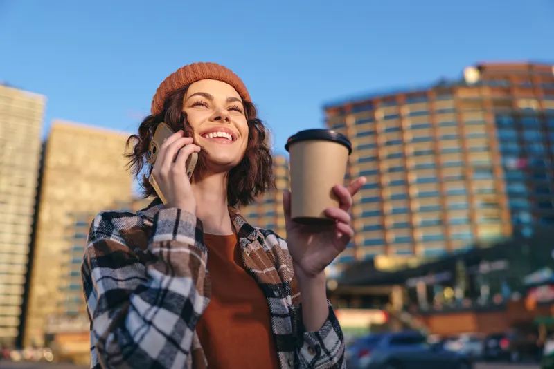 Woman holding coffee and phone in city, smile and beanie in warm sunlight during golden hour; candid lifestyle portrait emphasizing authenticity, mindful living and emotional storytelling.