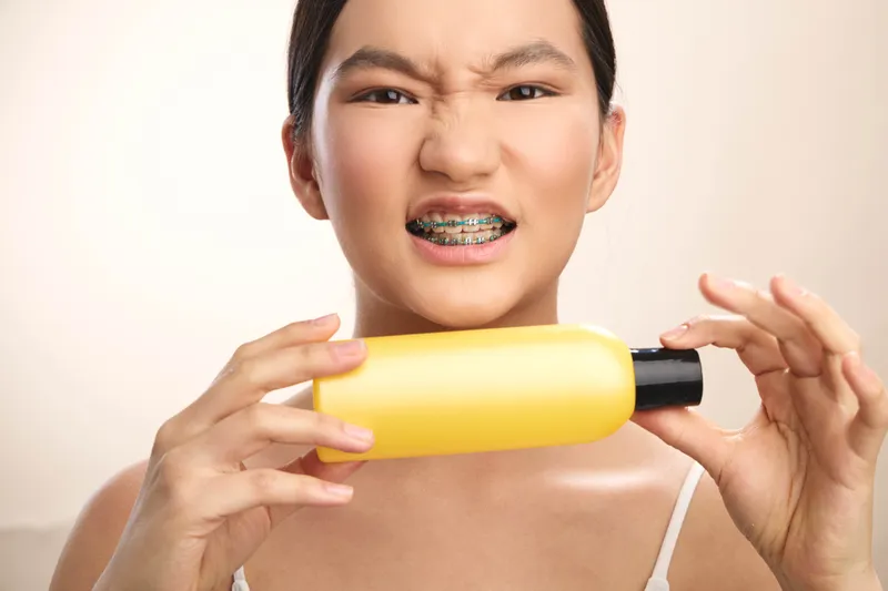 asian woman with braces is squeezing a yellow bottle at camera in a casual home setting, her expression playful and curious, creating a lighthearted and vibrant moment for a lifestyle photo.