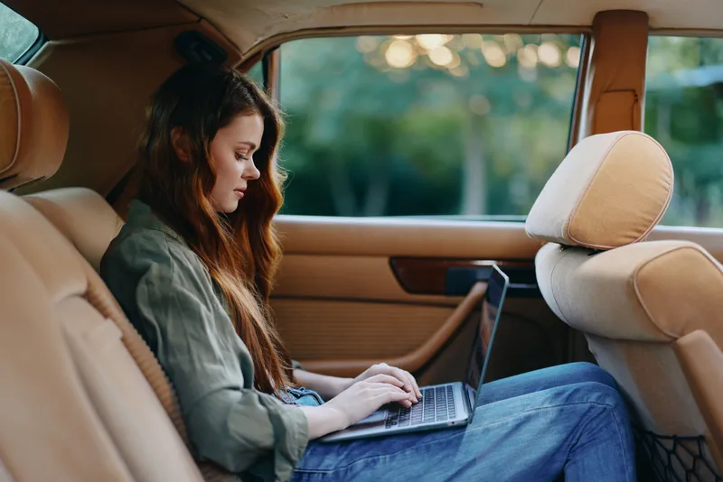 Young woman working on laptop in the backseat of a car, showcasing productivity and focus in a serene outdoor environment with a soft color palette.