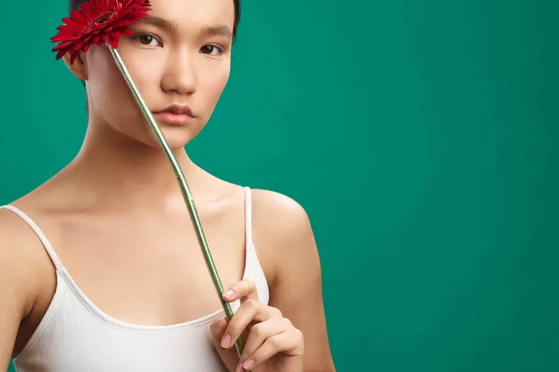 Asian woman holds a red flower near her face against a teal background, creating a calm, artistic portrait with soft lighting and minimal styling for a fresh mood.