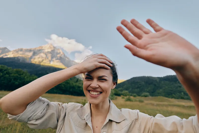 Smiling woman in a light shirt raises her hand in a sunny open field, enjoying a mountain backdrop and the carefree mood of a rural day.