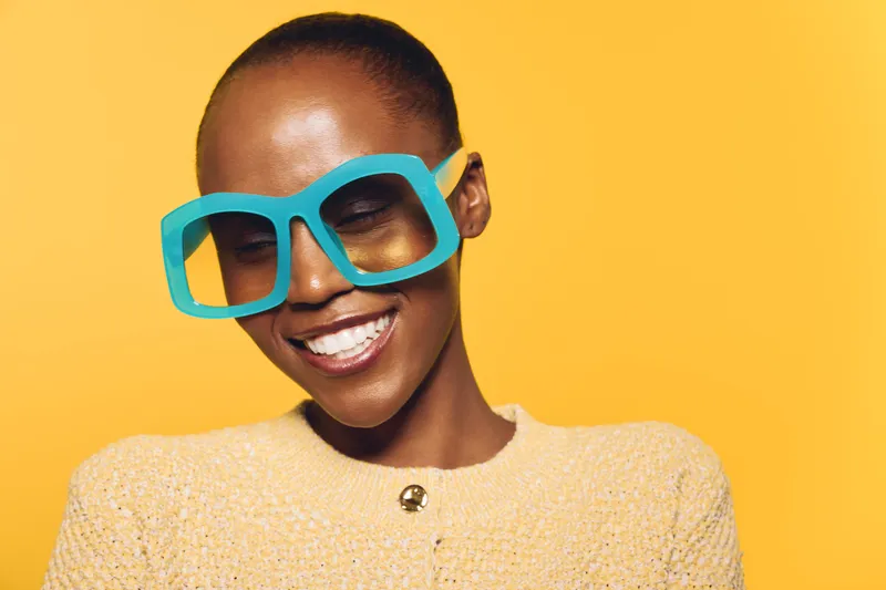 Smiling African woman with dark skin wearing large blue glasses and yellow sweater on yellow background. Concept of joy, fashion, beauty, and confident female portrait with bright colors.
