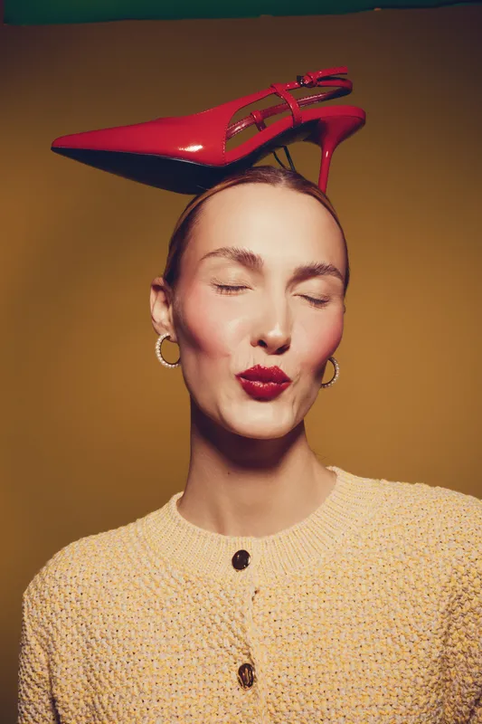 Woman with closed eyes and red lipstick balances red high heel shoe on head. She wears gold hoop earrings and beige knitted sweater. Concept of fashion, style, and playful elegance.