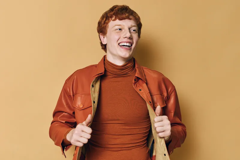 Young man with red hair smiling and posing wearing brown leather jacket and turtleneck on beige background for casual fashion and joyful lifestyle.