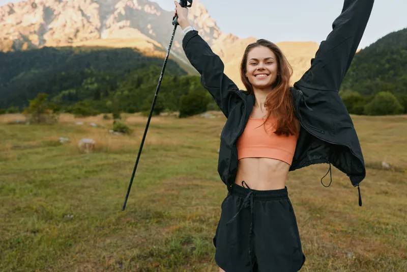 A cheerful young woman outdoors, wearing a cropped top and sporty jacket, raises her arms in triumph as she stands in a grassy field with distant mountains behind.