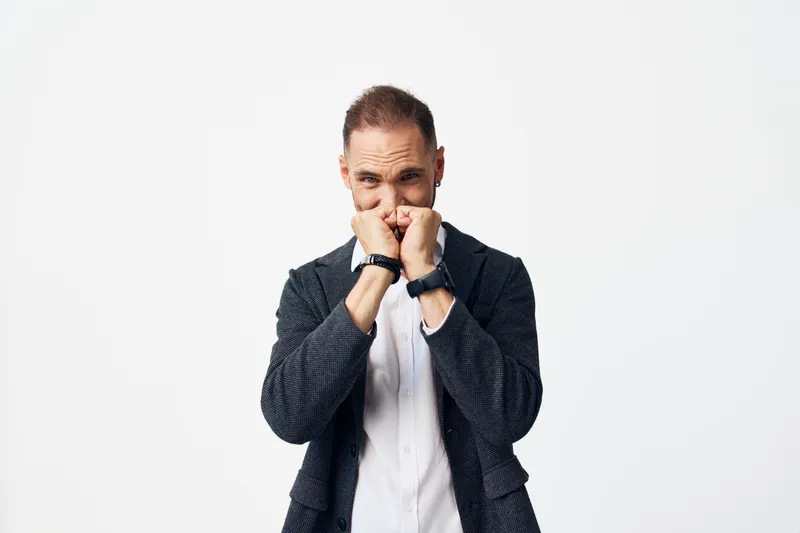 A professional business man stands against an isolated colored background, showing a tense emotion as he covers his face with his fists, signaling anticipation and focus.