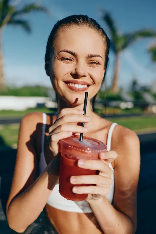 Vibrant outdoor portrait of a smiling woman enjoying a refreshing juice cup by a sunny tropical setting, capturing carefree summer lifestyle and relaxed poolside vibes.