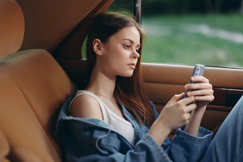Young woman in casual denim outfit sitting in a vintage car, focused on her smartphone in a serene outdoor setting, emphasizing relaxation and connectivity.