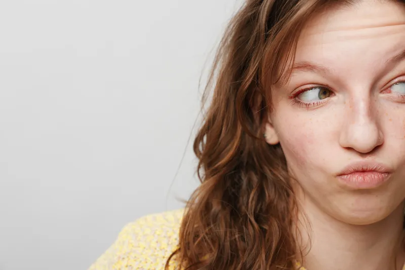 Close-up of a young woman with red hair making a skeptical face looking sideways. Neutral background highlights her natural expression and casual appearance.