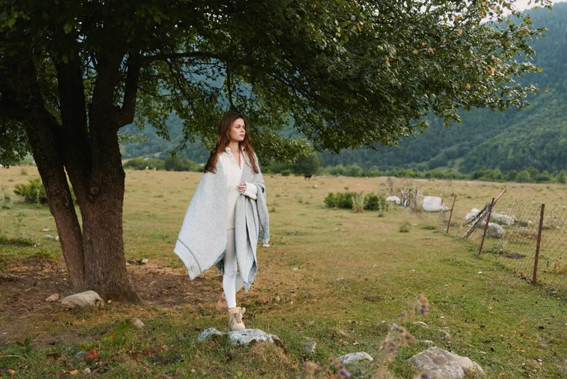 A solitary woman in a light shawl stands beside a tree in a grassy field, looking contemplative as distant hills rise behind her, creating a quiet mood.