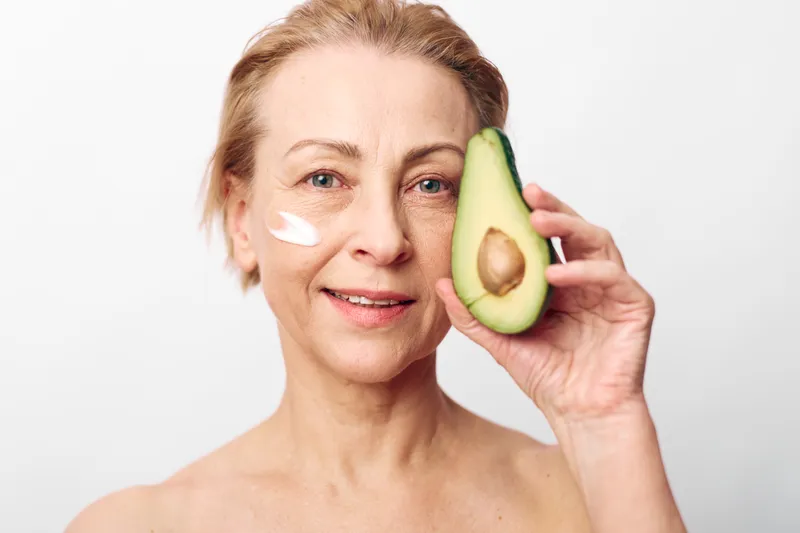 Mature woman with natural skin holds avocado half near face with cream on cheek. Concept of organic skincare, healthy lifestyle, and beauty wellness in natural light studio.