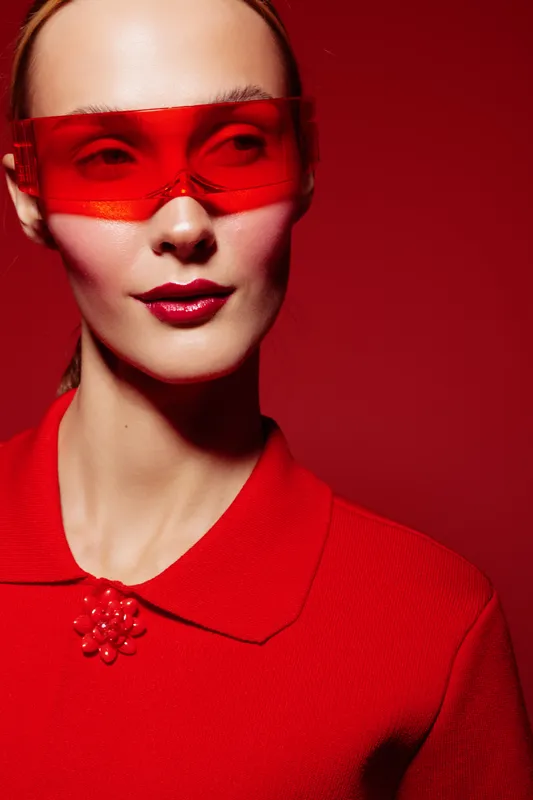 Stylish woman in red shirt and red translucent sunglasses poses on red background. Fashion portrait with bold makeup and modern accessories in vibrant monochrome style.