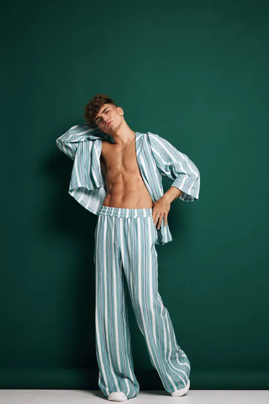 Fashion model posing in striped pajama set with open shirt revealing muscular torso over green background. Casual men sleepwear, relaxed style with confident stance and soft lighting for clothing