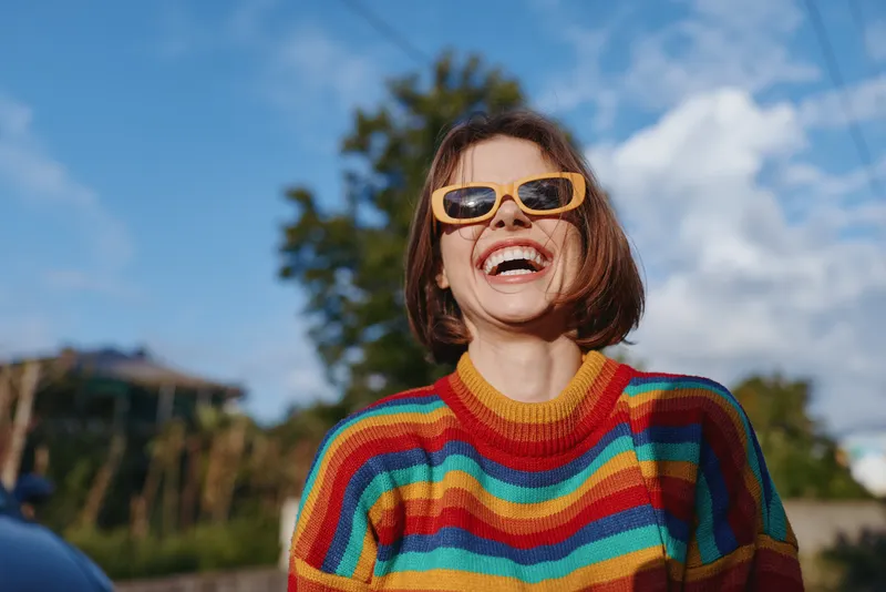 Woman smiling in a colorful striped sweater and yellow sunglasses outdoors, young adult laughing and looking up under a sunny blue sky, casual outfit lifestyle travel portrait.
