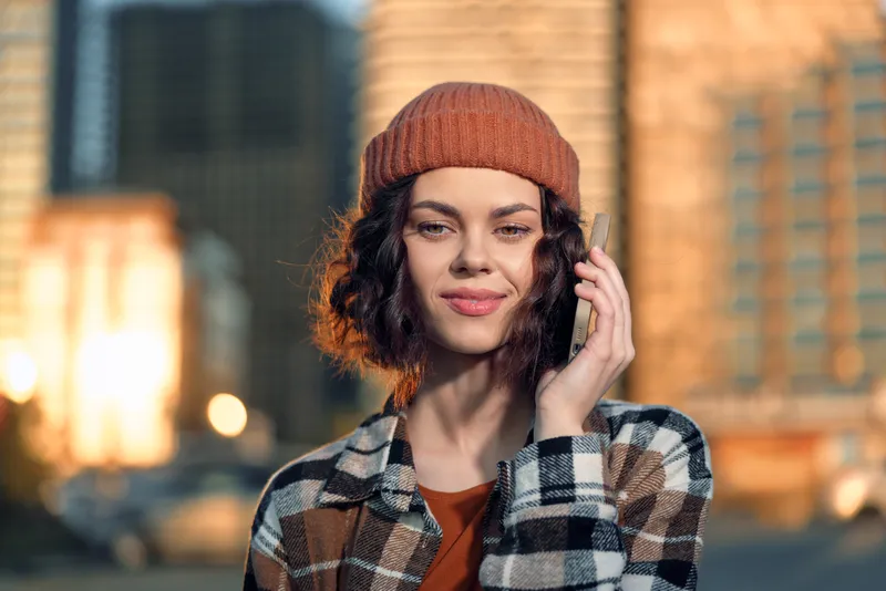 woman phone city portrait smile beanie candid authenticity golden Young woman in beanie talking on phone against city skyline at golden hour, candid lifestyle, mindful living and emotional