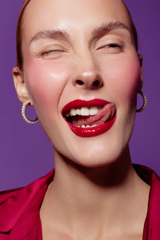 Close-up of a woman with red lipstick winking and licking lips against purple background. Concept of beauty, makeup, facial expression, and confident female model in studio lighting.