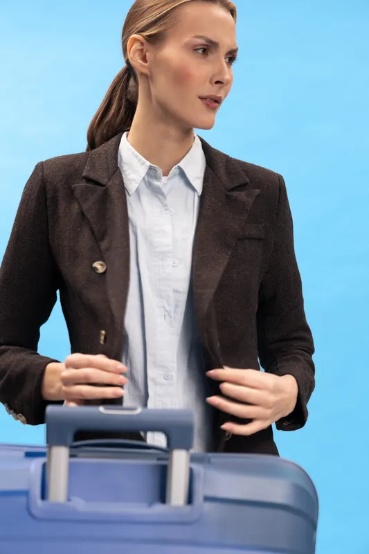 Young businesswoman in brown blazer stands behind blue suitcase against blue background. Travel concept with professional woman preparing for business trip or journey in studio lighting.