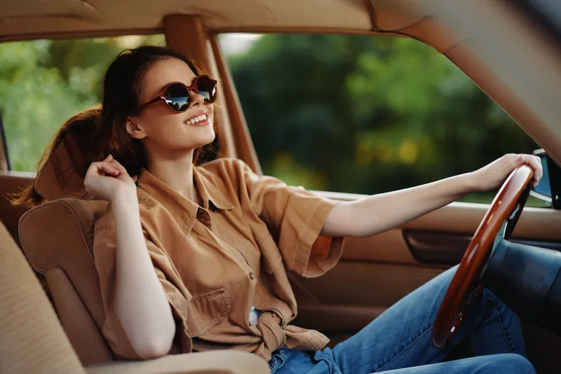 Young woman in stylish sunglasses enjoying a ride in a vintage car, embodying carefree summer vibes and happiness in a lush green setting.