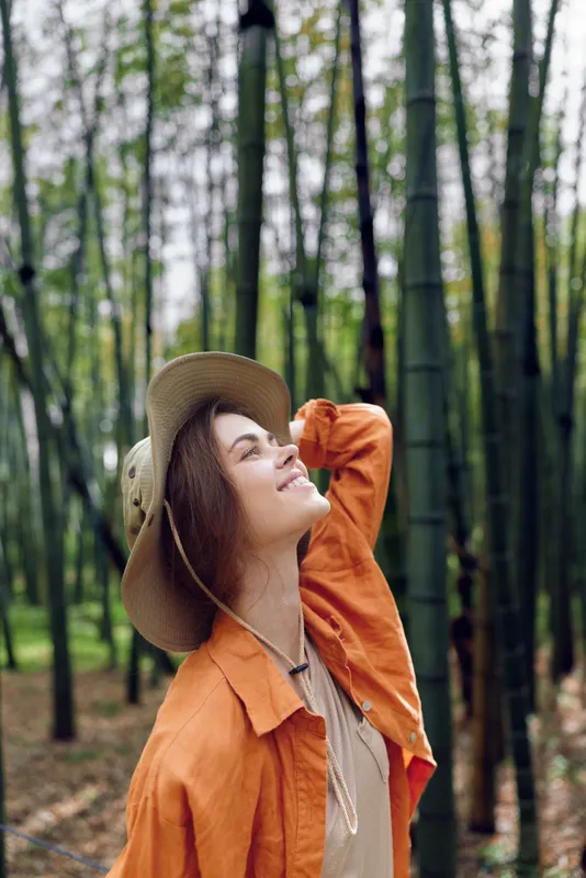 Woman in hat smiling portrait in a bamboo forest, outdoor nature scene with green foliage and sunlight, relaxed travel lifestyle, casual fashion and peaceful exploration.