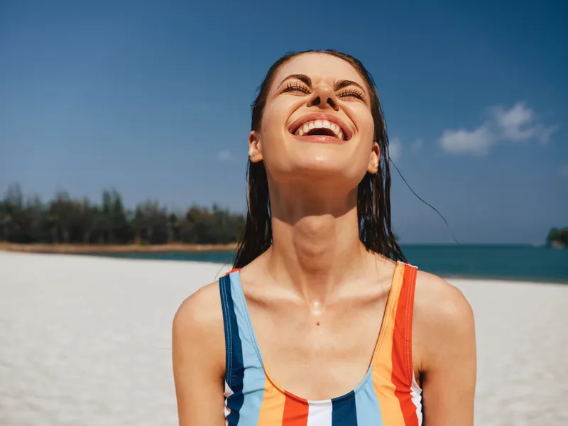 Close up of a joyful woman on a sunny beach, smiling with head tilted back, wearing a colorful striped swimsuit, while a blue sky and calm sea form a vibrant backdrop.