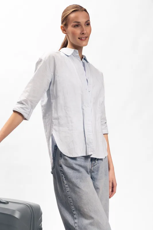 Young woman in casual white shirt and jeans stands with suitcase on white background. Travel concept with relaxed female traveler ready for trip, isolated in bright studio lighting.